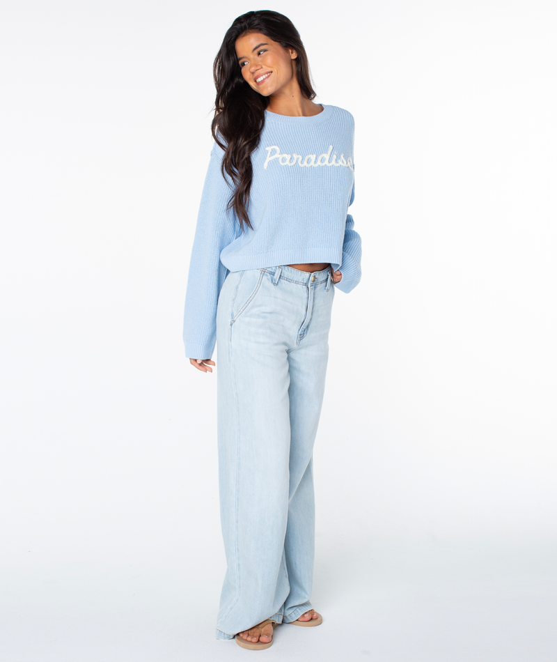 Woman wearing a Roxy Swell Season Sweater in Cerulean Paradise, wide-leg light wash jeans, and sandals, standing and smiling against a plain white background.