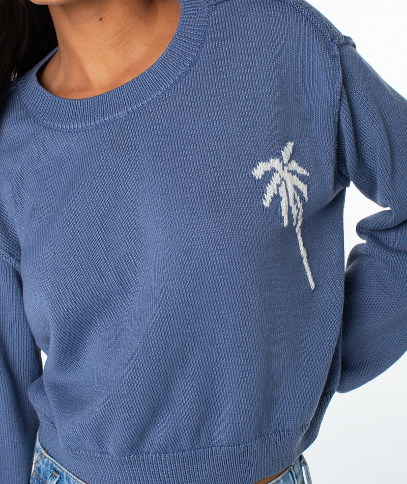 A person in the Roxy Swell Season Sweater—a blue knit with a white palm tree design—paired with light blue jeans. Photo is cropped at the shoulders and waist.