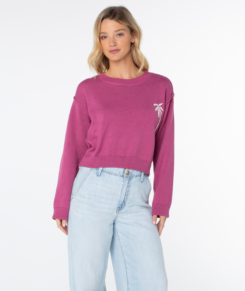 A woman wears the Roxy Swell Season Sweater in magenta with a small palm tree on the chest and light blue high-waisted jeans, posing against a plain white background.