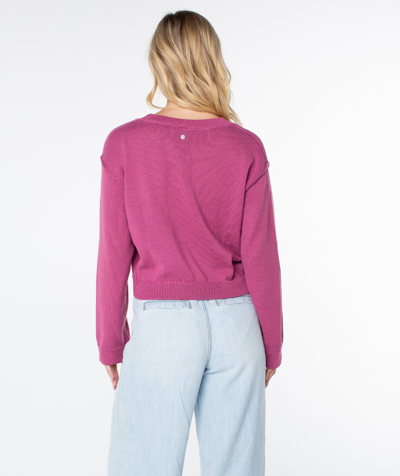 A person with long blonde hair is wearing the Roxy Swell Season Sweater in magenta and light blue jeans, facing away from the camera against a plain white background.