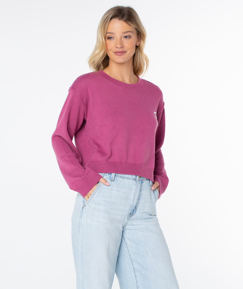 A woman in a magenta Roxy Swell Season Sweater and light blue high-waisted jeans stands with her hands in her pockets, smiling slightly against a white background.