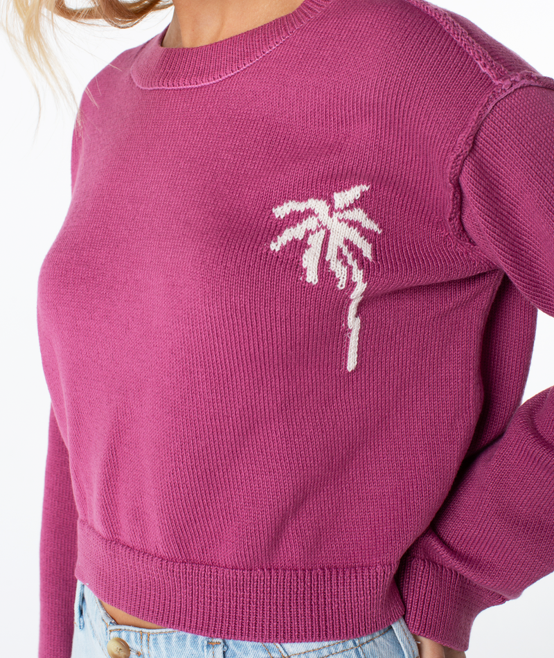 A person wearing a magenta Roxy Swell Season Sweater with a white palm tree design on the left chest and light blue jeans.