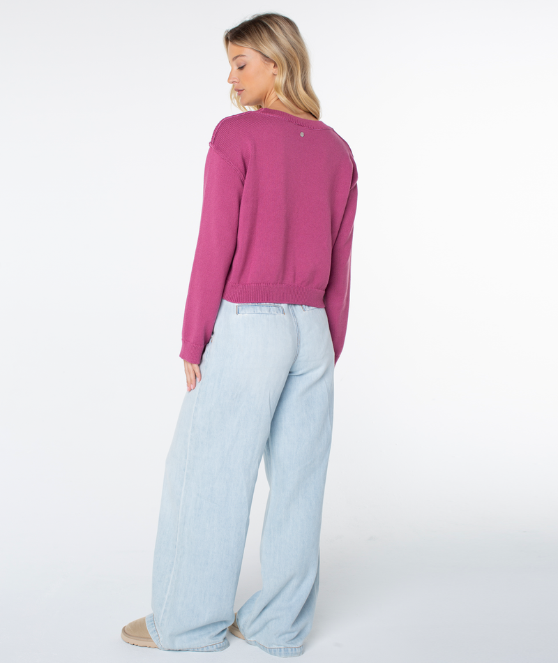 A woman with long blonde hair wears the Roxy Swell Season Sweater in magenta and light blue wide-leg jeans, standing slightly turned away against a plain white background.