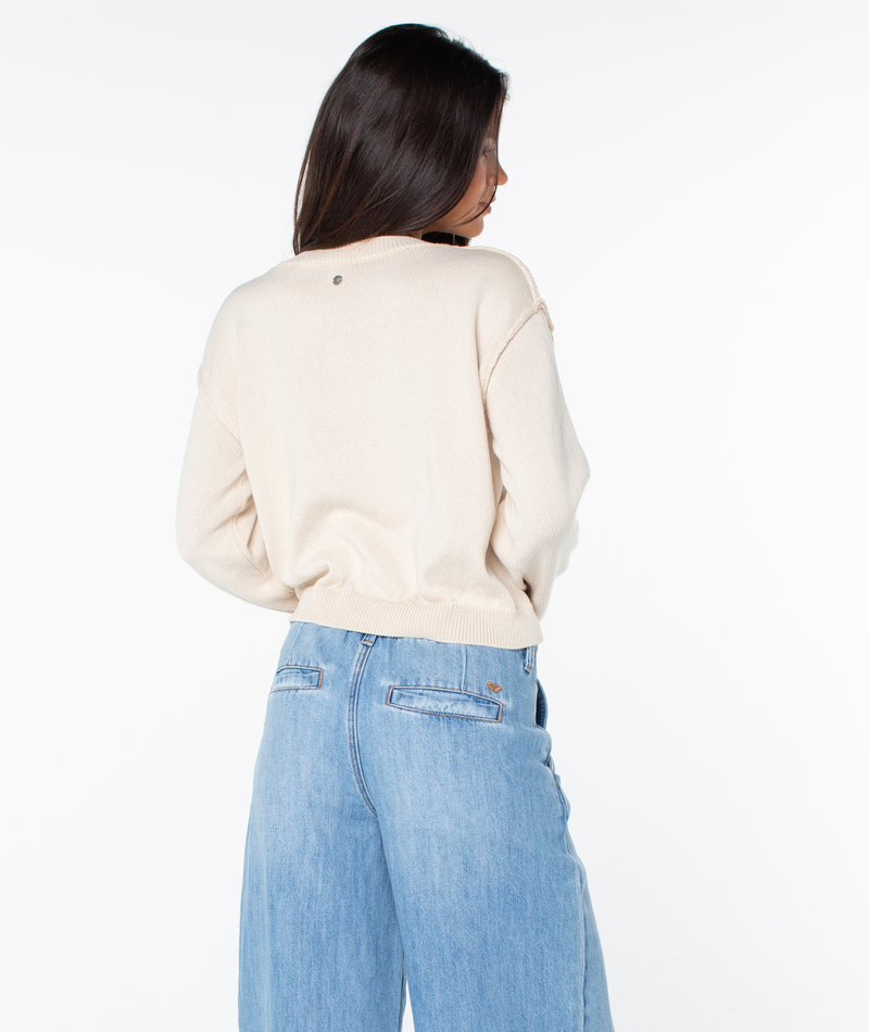 A woman with long dark hair wears the Roxy Swell Season Sweater in light beige and high-waisted light blue jeans, standing against a plain white background.