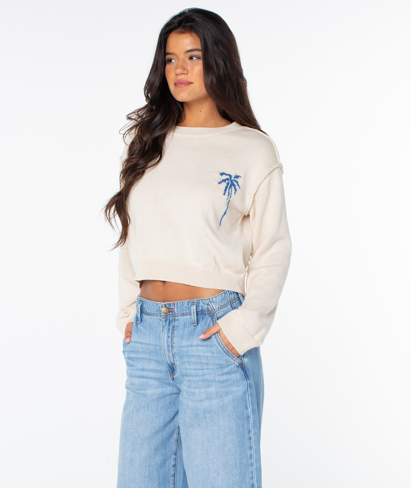 A woman in a cream Roxy Swell Season Sweater with a blue palm tree design and light blue jeans stands with hands in pockets against a white background.