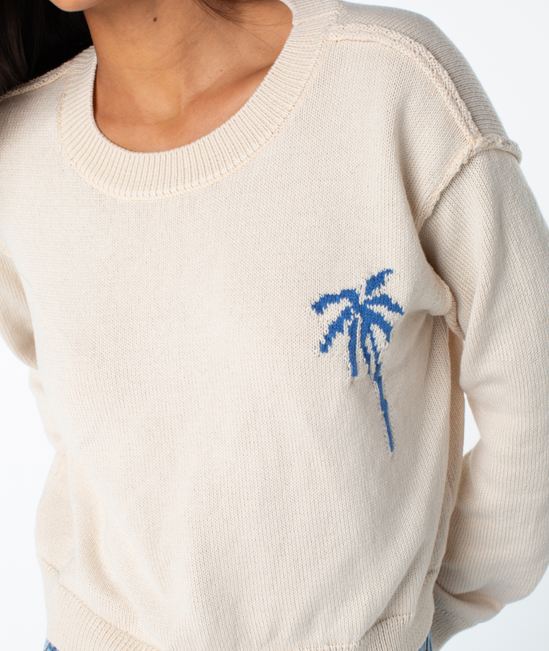 A person wearing the Roxy Swell Season Sweater in cream with a blue palm tree design on the chest, photographed from shoulders to hips with partially visible hair.