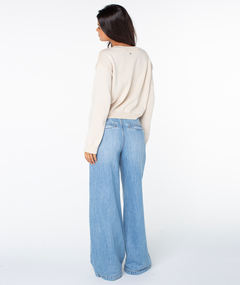 A woman with long dark hair stands against a white background, facing away, wearing the cream-colored Swell Season Sweater by Roxy and light blue wide-leg jeans, her left hand resting by her side.