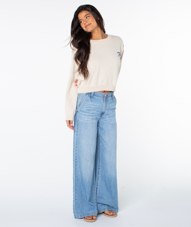 A woman with long dark hair wears the Swell Season Sweater by Roxy in Parchment Palm Tree and high-waisted wide-leg blue jeans, standing against a white background and smiling slightly to the side.