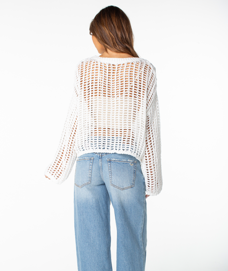 A woman faces away in light blue jeans and the Roxy Lost At Sea Long Sleeve Top, a white open-knit layer over a beige shirt, set against a plain white background.