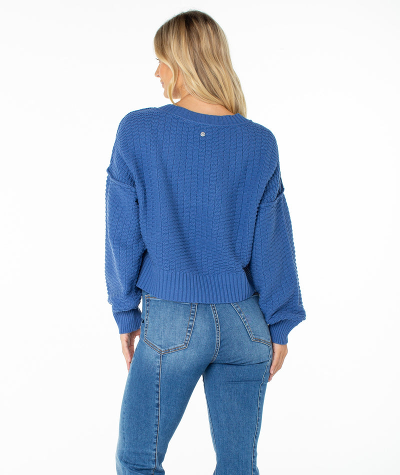 A woman with blonde hair wears the Roxy Study Hall Sweater, a cozy knit blue design, paired with blue jeans as she stands with her back to the camera against a plain white background.