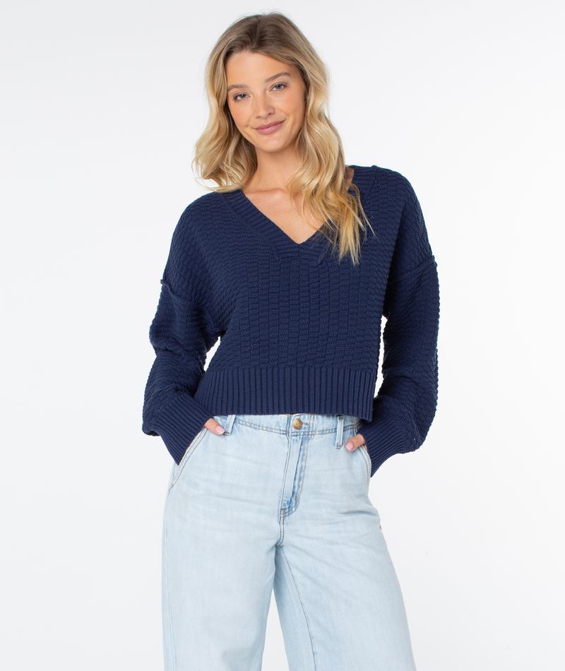 A person wearing a dark blue, cozy knit V-neck Study Hall Sweater by Roxy with a textured pattern and light wash denim jeans.