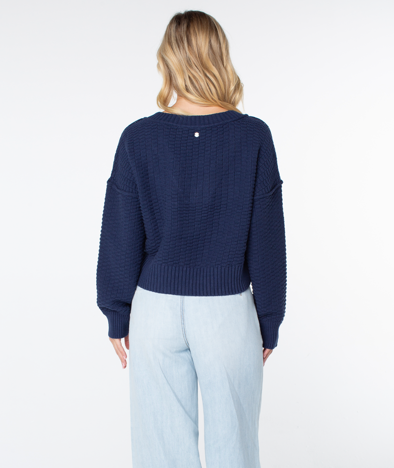 Back view of a person wearing a cozy navy blue textured Roxy Study Hall Sweater with a ribbed hem and cuffs, paired with light-wash jeans.
