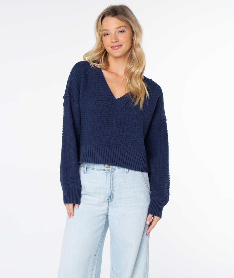 A woman wears a cozy, navy blue Roxy Study Hall Sweater featuring a V-neck and textured knit pattern, paired with light-wash denim jeans.