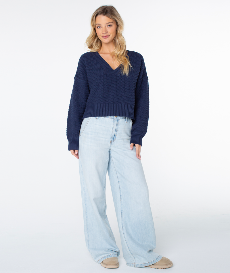 A person wearing a cozy, dark blue, textured Study Hall Sweater by Roxy with drop shoulders, paired with light-wash, wide-leg jeans and tan slip-on shoes.