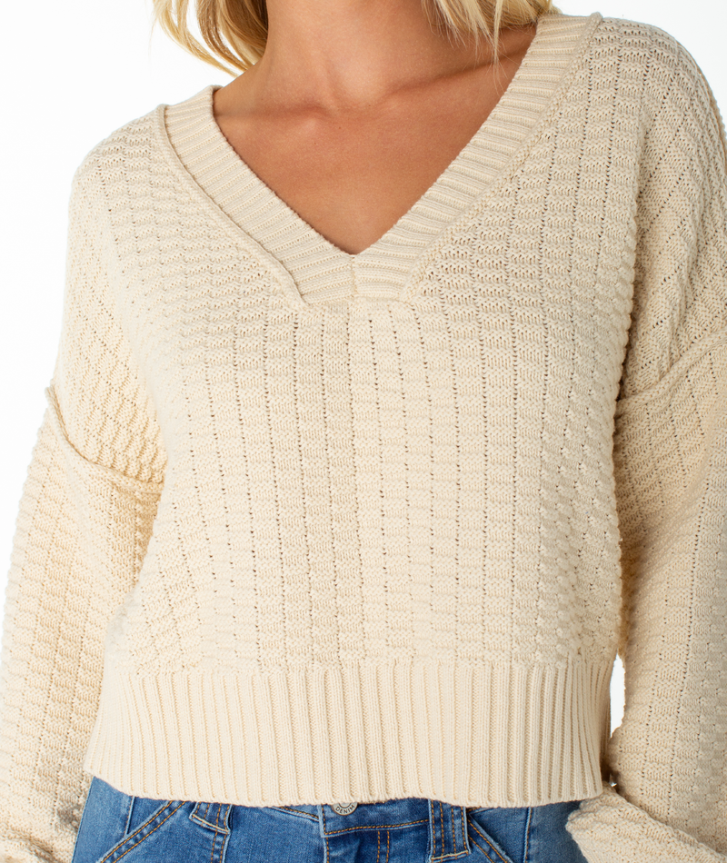 A person wearing the Roxy Study Hall Sweater—a cream-colored, cozy knit V-neck with long sleeves—paired with blue jeans. The photo is cropped at the neck and hips.