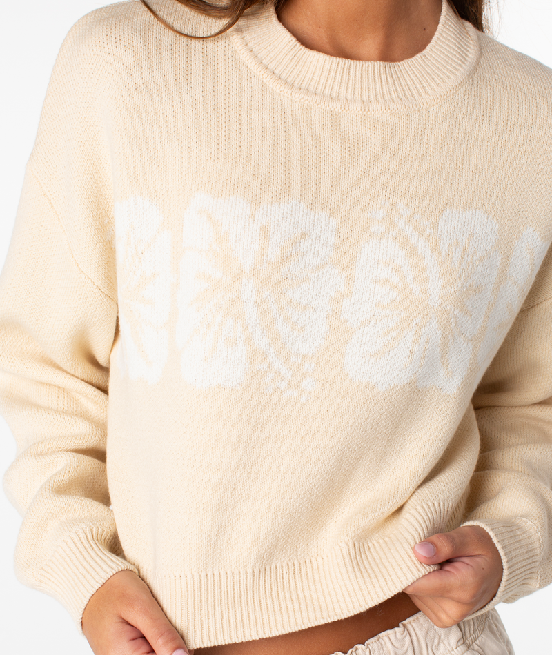 A person wears the Roxy Hibiscus Harmony Sweater—a cream-colored, cozy cotton-blend piece featuring white hibiscus flower designs across the chest—paired with light-colored pants. Only their lower face and upper body are visible.
