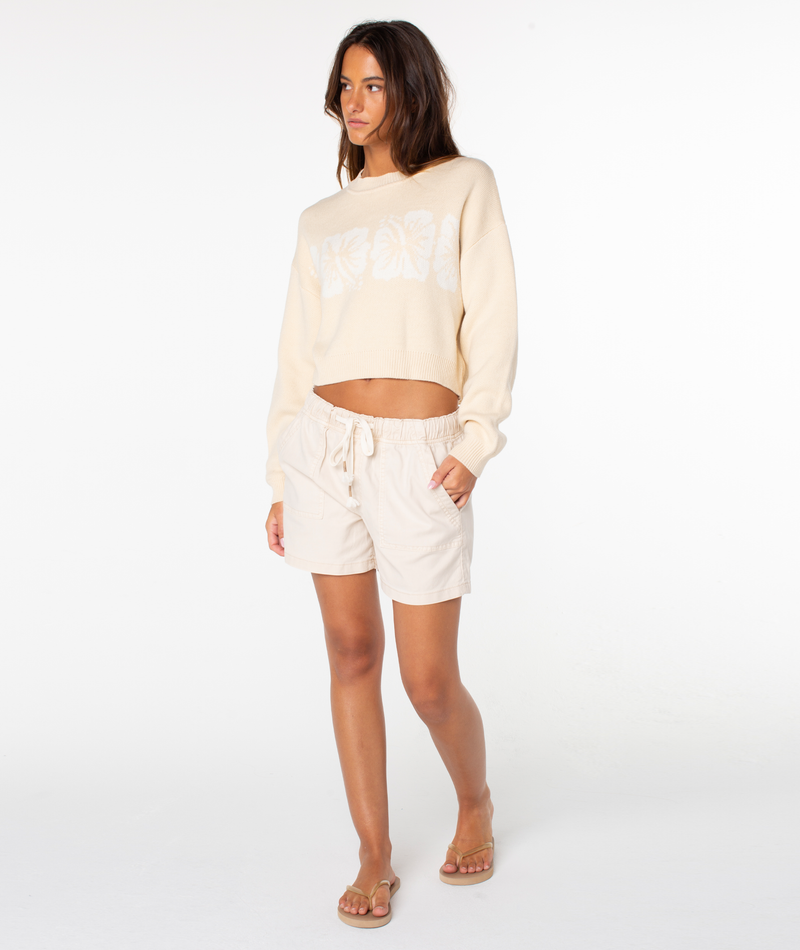 A woman stands in the Roxy Hibiscus Harmony Sweater—a cream, relaxed-fit cotton blend with a white floral design—paired with beige drawstring shorts and tan sandals against a plain white background.
