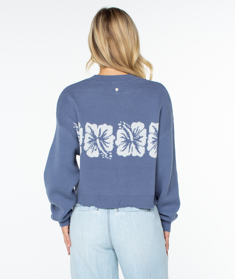 A person with long blonde hair, wearing the Roxy Hibiscus Harmony Sweater in blue with a white hibiscus pattern on the back and light blue jeans, stands facing away from the camera.