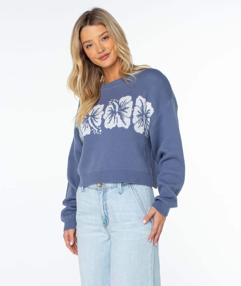 A woman with long blonde hair wears the Roxy Hibiscus Harmony Sweater—a cozy cotton-blend blue top with large white flowers—paired with light blue jeans, set against a plain white background. Perfect for breezy beach nights.