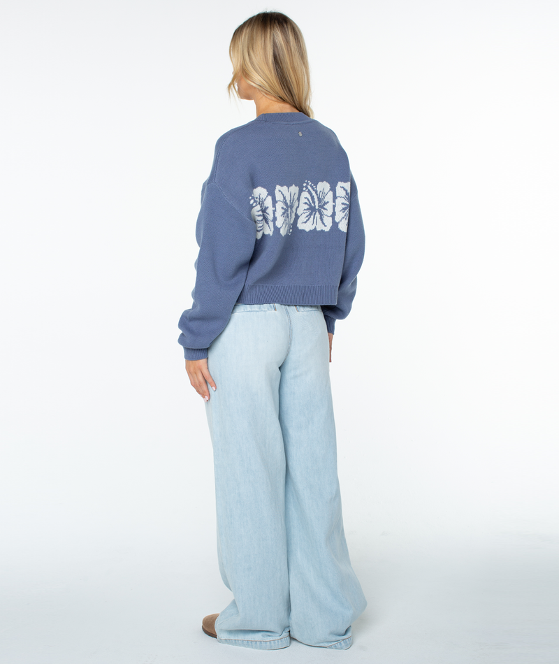 A person faces away, wearing the relaxed fit Hibiscus Harmony Sweater by Roxy—blue with white butterfly designs—and loose, light blue jeans. Blond hair falls against a plain white background. Perfect for breezy beach nights.