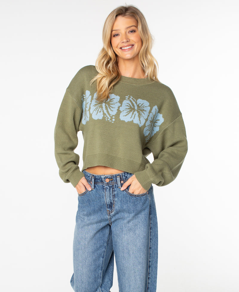 A woman stands facing the camera against a plain white background, smiling with her hands in her pockets. She is wearing blue jeans and the Roxy Hibiscus Harmony Sweater—a cozy green cropped cotton blend featuring blue floral designs.