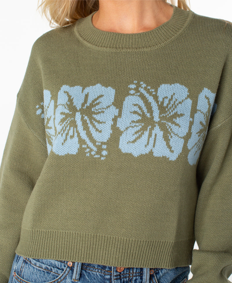 A person wears the Roxy Hibiscus Harmony Sweater, featuring an olive green shade with light blue hibiscus flower patterns across the chest, crafted from a cozy cotton blend for ultimate comfort, paired with blue jeans.