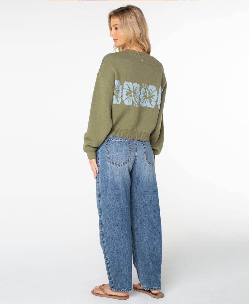 A woman with blonde hair stands sideways against a plain white background, wearing the Roxy Hibiscus Harmony Sweater—an olive cotton blend sweater featuring blue floral designs on the back—paired with relaxed fit blue jeans and flip-flops.