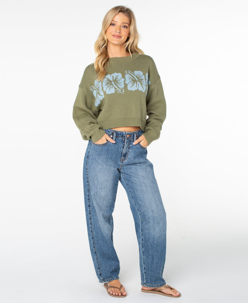 A woman with long blonde hair wears the Roxy Hibiscus Harmony Sweater in Oil Green Hibiscus, a cozy cotton-blend sweater featuring blue floral designs. She pairs it with relaxed-fit blue wide-leg jeans and sandals, smiling with one hand on her hip against a plain white background.