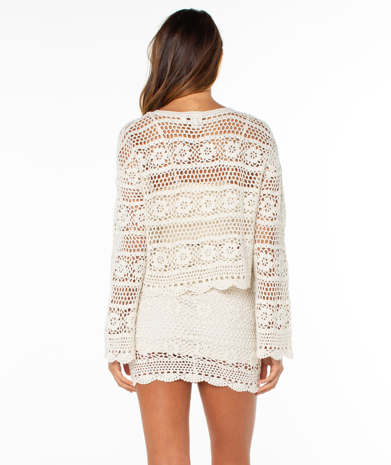 A woman stands facing away, wearing the Blue Water Sweater by Roxy. The cream-colored top features intricate patterns and scalloped edges; her long hair is loose, and the background is plain white.