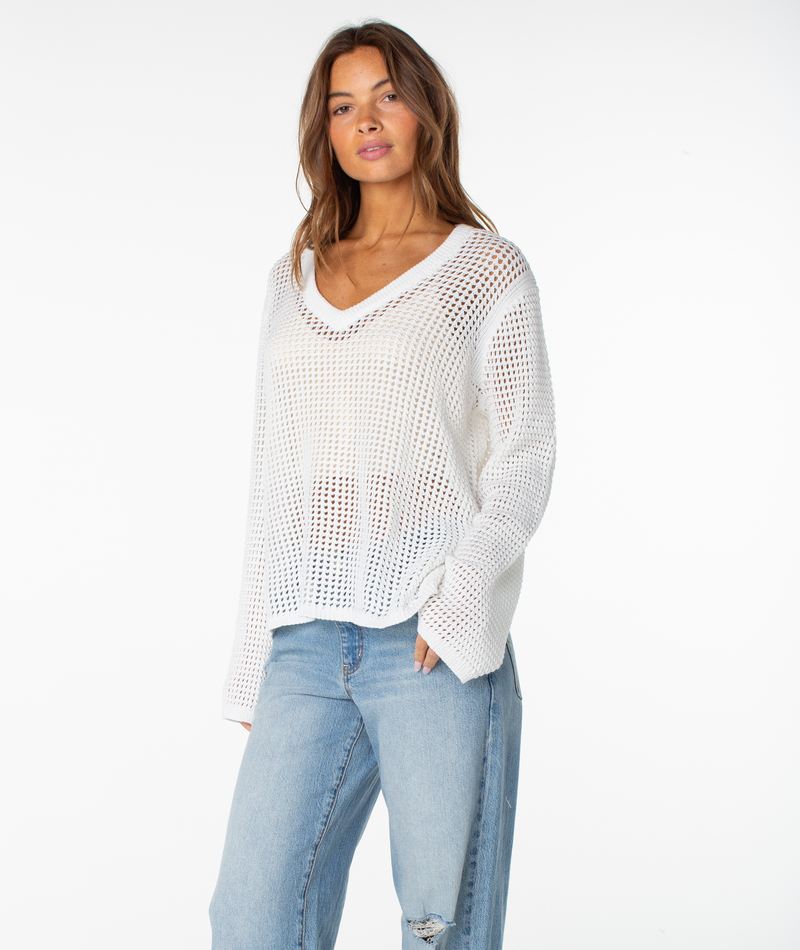 A woman stands against a white background wearing the Roxy Summer Nights V Neck Tunic over a white top and light blue jeans, hand in her pocket and looking relaxed—an ideal summer staple.