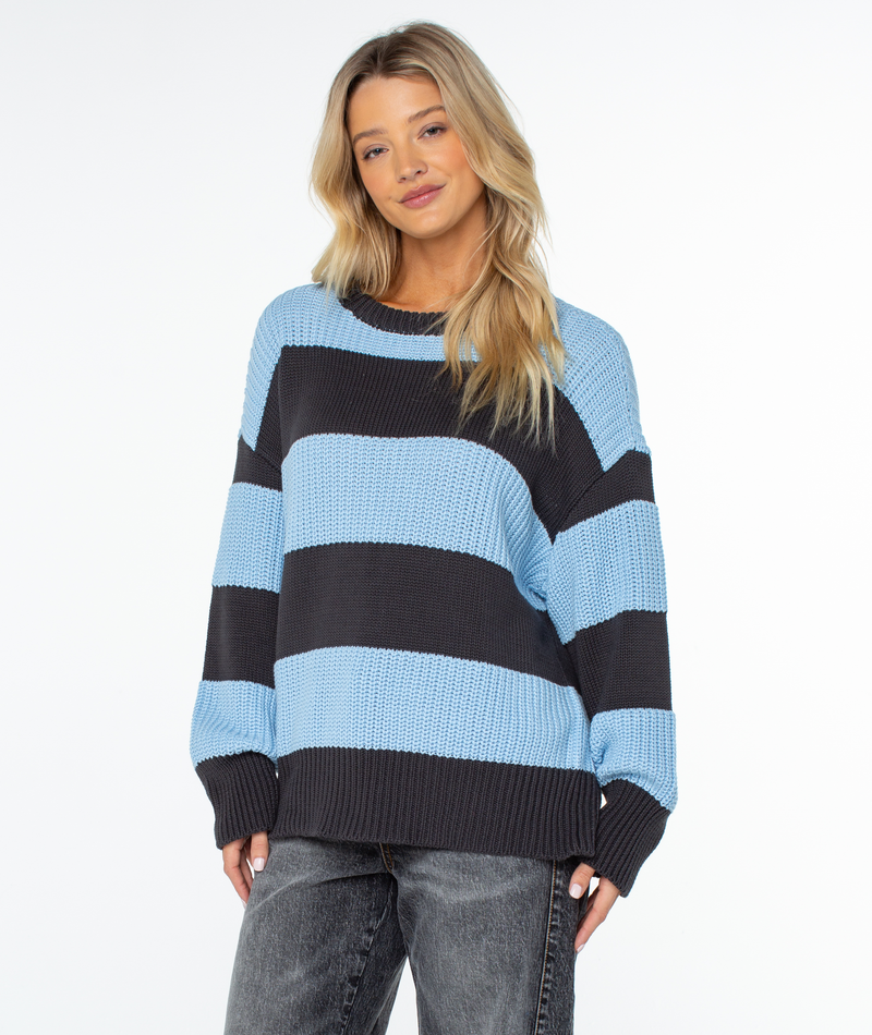 A person wearing a Roxy West Side Sweater featuring an oversized chunky knit design with wide horizontal light blue and dark gray stripes, paired with dark denim jeans.