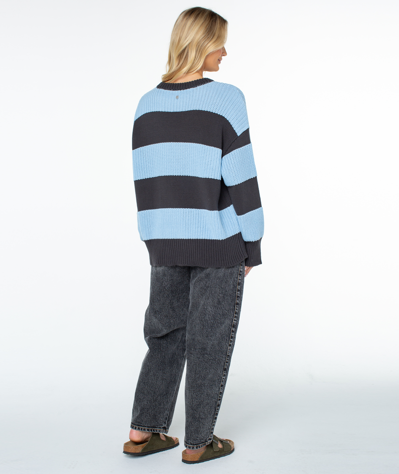A person viewed from behind wearing a Roxy West Side Sweater in an oversized, chunky knit with light blue and dark gray stripes, dark gray jeans, and olive green slide sandals.