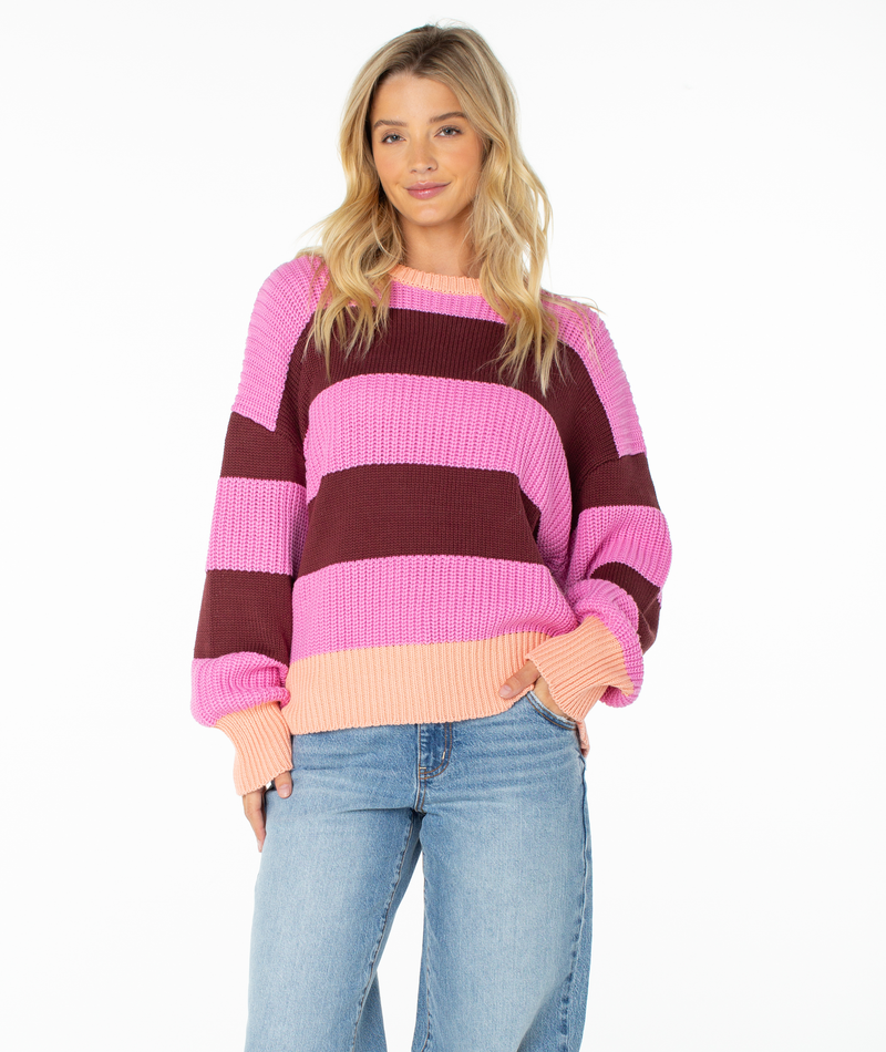 A woman with long blonde hair stands against a white background, exuding relaxed confidence in the Roxy West Side Sweater. The sweater features bold fuchsia pink stripes and a boxy drape that complements her effortlessly stylish look. She pairs it with light blue jeans for a cozy, on-trend outfit.