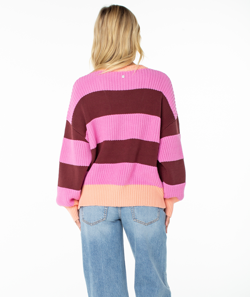 A person with long blonde hair stands with their back to the camera against a white background, wearing the Roxy West Side Sweater—an oversized chunky knit in pink and burgundy stripes with wide sleeves—paired with light blue jeans.