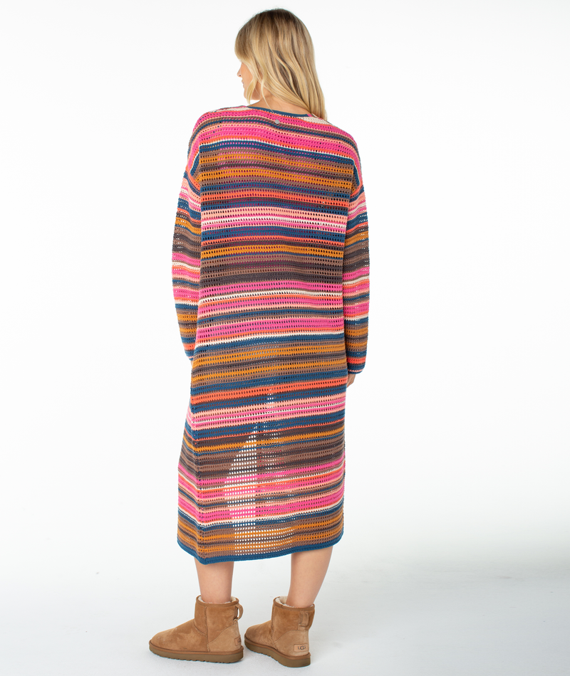 A person stands facing away, wearing the Roxy Sea Wind Cardigan—a long, multicolored knit piece featuring pink, orange, brown, and blue horizontal stripes reminiscent of a classic cotton blend. They pair it with tan boots against a plain white background.