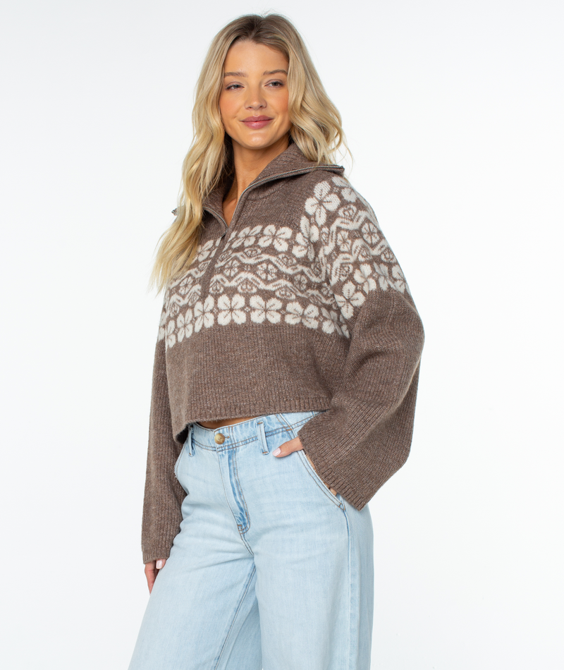 A woman wears a brown, relaxed-fit, cropped Roxy Cedar Grove Sweater with a cream-colored floral pattern across the chest and sleeves, paired with light-wash jeans.