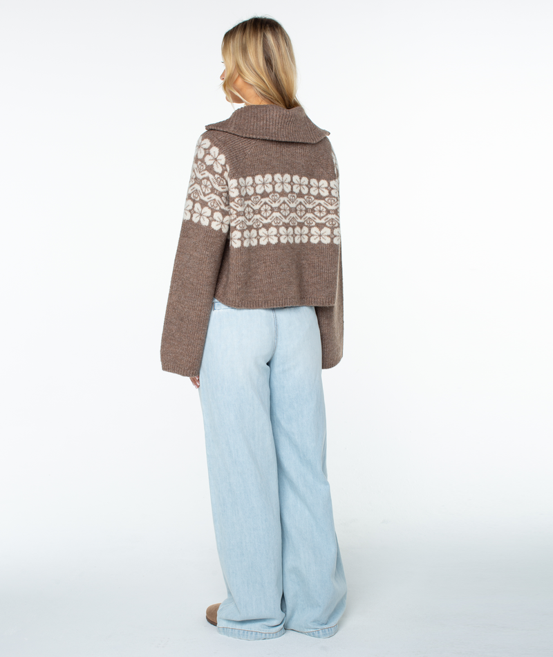 A person viewed from the back, wearing a brown, cropped, floral-patterned Roxy Cedar Grove Sweater with relaxed-fit, light-wash jeans.
