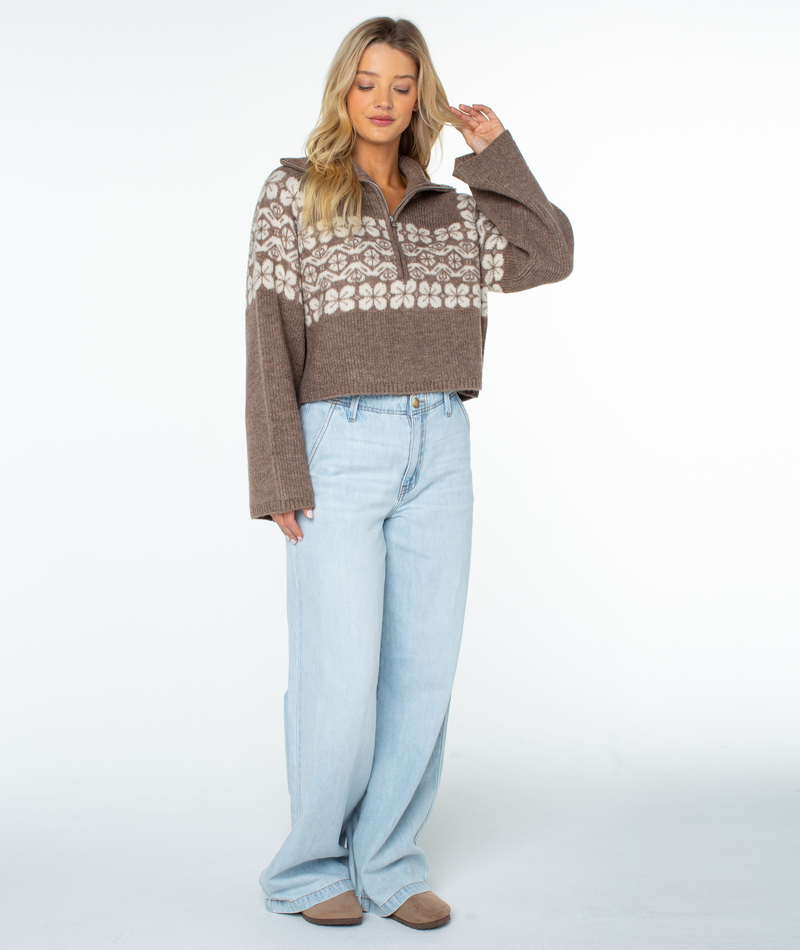 A woman wearing a brown Roxy Cedar Grove Sweater, a relaxed, cropped quarter-zip with a white floral pattern across the chest, paired with light-wash wide-leg jeans.