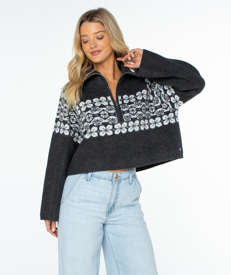 A person wears a Roxy Cedar Grove Sweater, a dark gray, cropped quarter-zip with a relaxed fit and white repeating floral pattern across the chest and shoulders, paired with light-wash jeans.