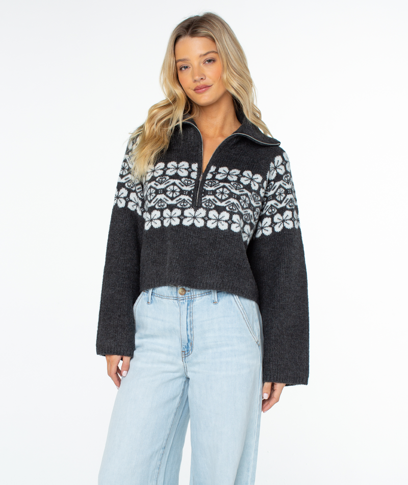 A woman wears a dark gray, cropped, relaxed-fit Roxy Cedar Grove sweater featuring a white snowflake pattern across the chest and light-wash jeans, exuding wintery vibes.