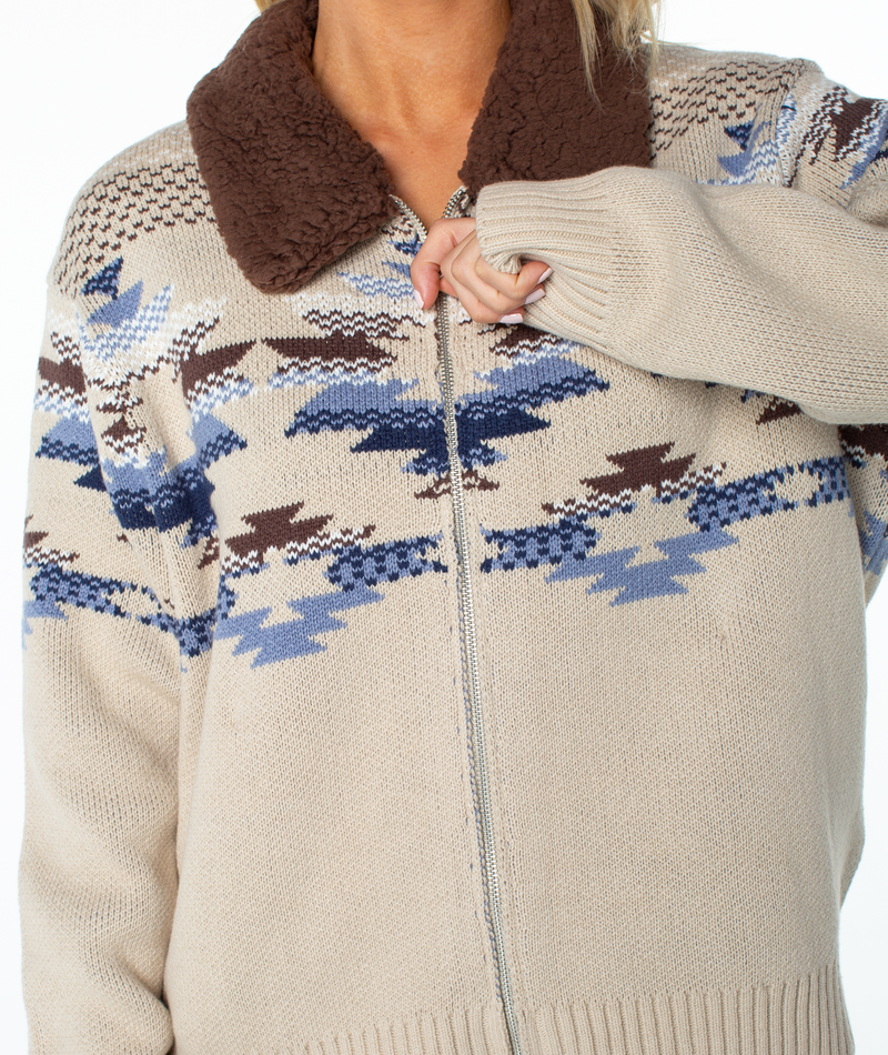 The Roxy Wildwood Sweater is a beige zip-up knit sweater featuring a brown faux-shearling collar and a blue and brown geometric statement print; a versatile cold-weather staple.