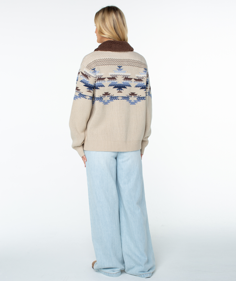 Back view of a person wearing a beige Roxy Wildwood Sweater featuring a blue and brown geometric pattern and a brown collar, paired with light-wash wide-leg jeans.