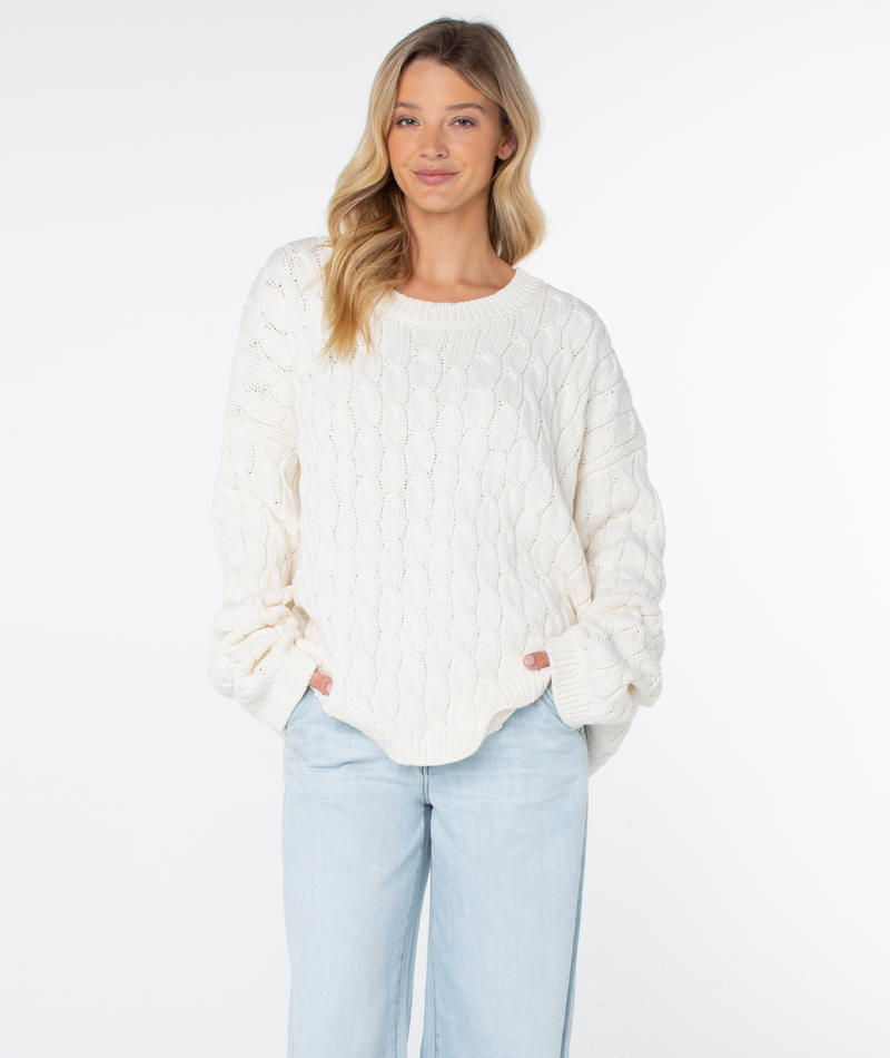 A woman wears a cozy Roxy Cold Front Cable Sweater with a round neckline and light-wash jeans.