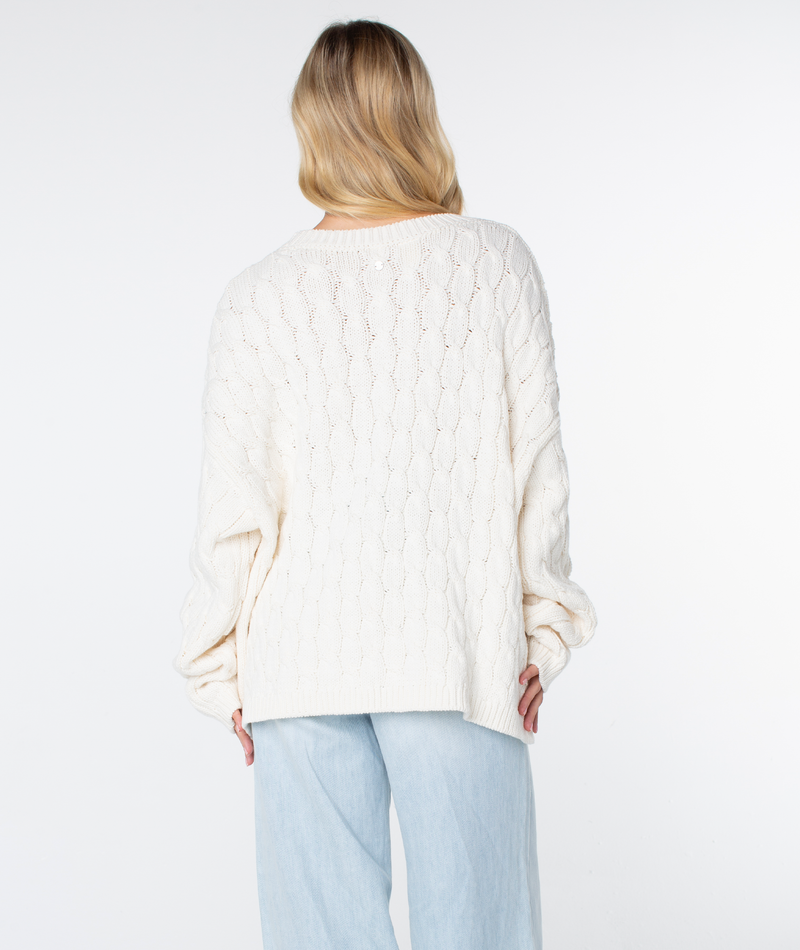 Back view of a Roxy Cold Front Cable Sweater in cream, featuring a cozy long-sleeve cable-knit design worn with light-wash jeans.