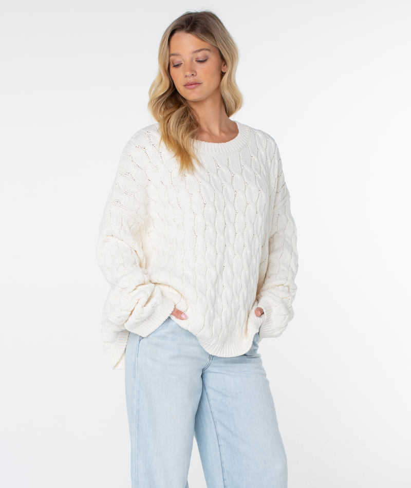 A woman wearing an oversized, cozy Roxy Cold Front Cable Sweater with long sleeves and light-wash jeans.