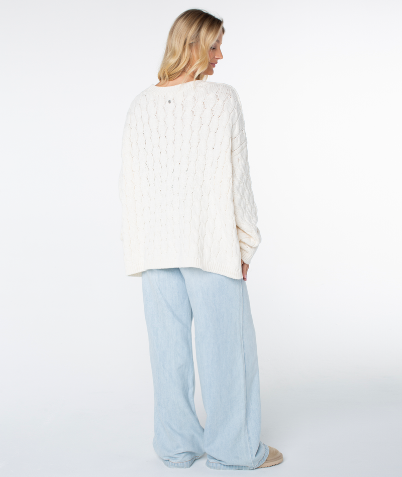 A person seen from behind, wearing a cozy, cream-colored Roxy Cold Front Cable Sweater and wide-leg light-wash jeans.