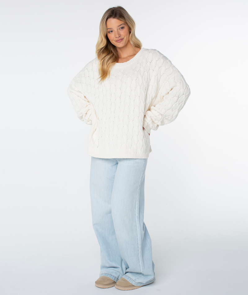 A person wears a cozy, coconut milk solid Roxy Cold Front Cable Sweater with drop shoulders paired with loose-fitting light-wash jeans.