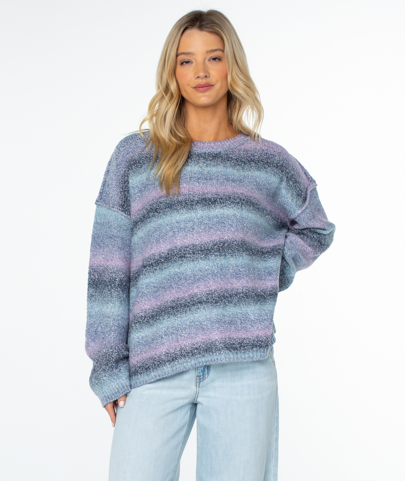 Woman wearing a Roxy Cold Front Sweater in Cerulean Stripe, featuring a relaxed fit, long-sleeved crewneck design with horizontal stripes in shades of blue and purple.