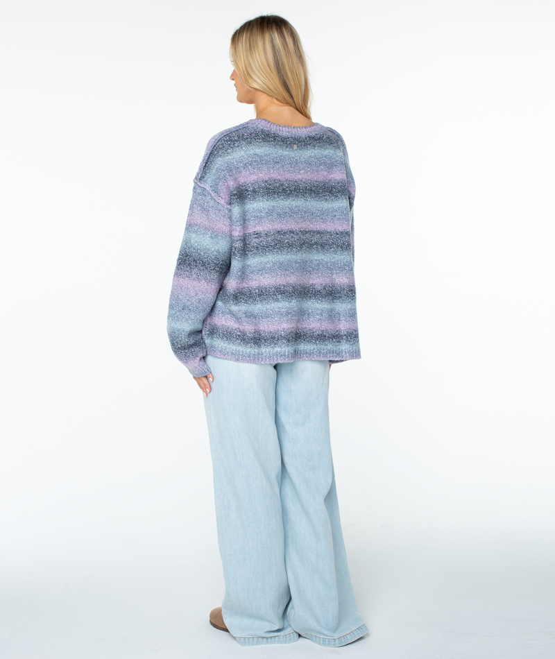 A person seen from behind wears the Roxy Cold Front Sweater, a slouchy, loose-fitting, long-sleeved top with blue and purple horizontal stripes, paired with light-wash wide-leg jeans.