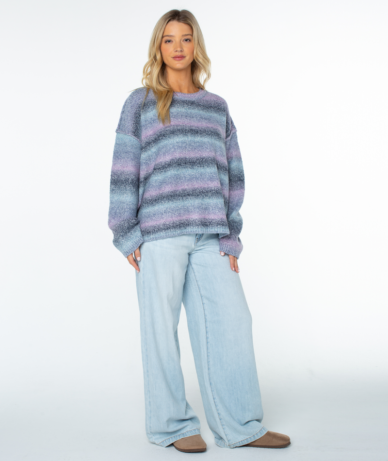 A woman wears a Roxy Cold Front Sweater in relaxed-fit horizontal stripes of blue and purple, paired with light-wash wide-leg jeans.